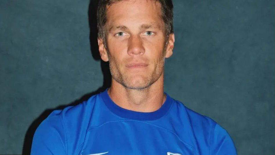 A handout picture of Tom Brady wearing a blue sports top. He is standing against a grey background and looking straight at the camera.