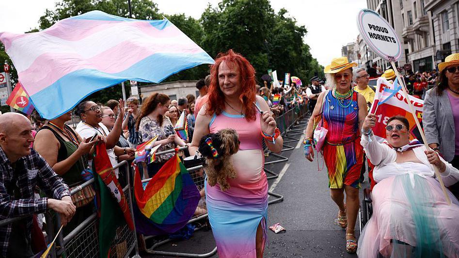 Three trans people walk past a series of crowd control barriers as people watch, cheer and wave flags. 
