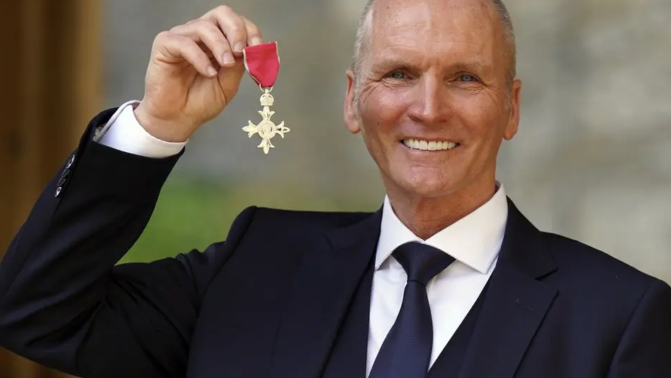 Thomas is holding is MBE in 2021. He is wearing a dark suit and tie and smiling at the camera.