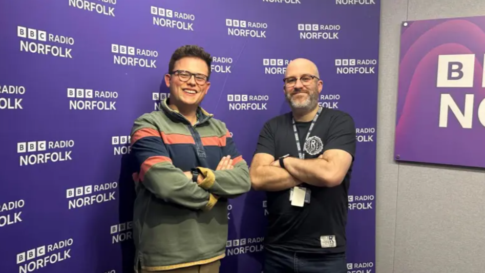 Two men stand with their arms crossed as they pose for the camera in front of a purple signage board with dozens of the same logo for BBC Radio Norfolk.