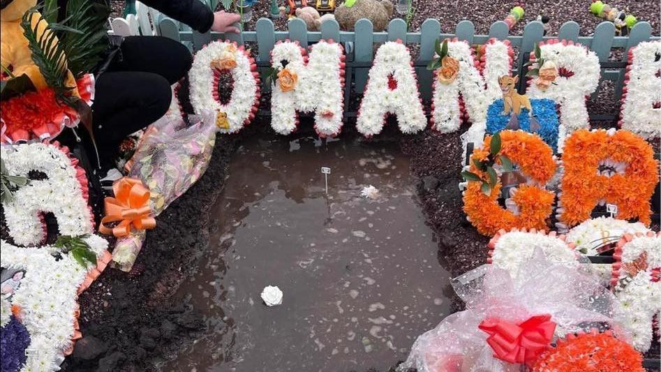 A flooded grave with orange and white floral tributes.