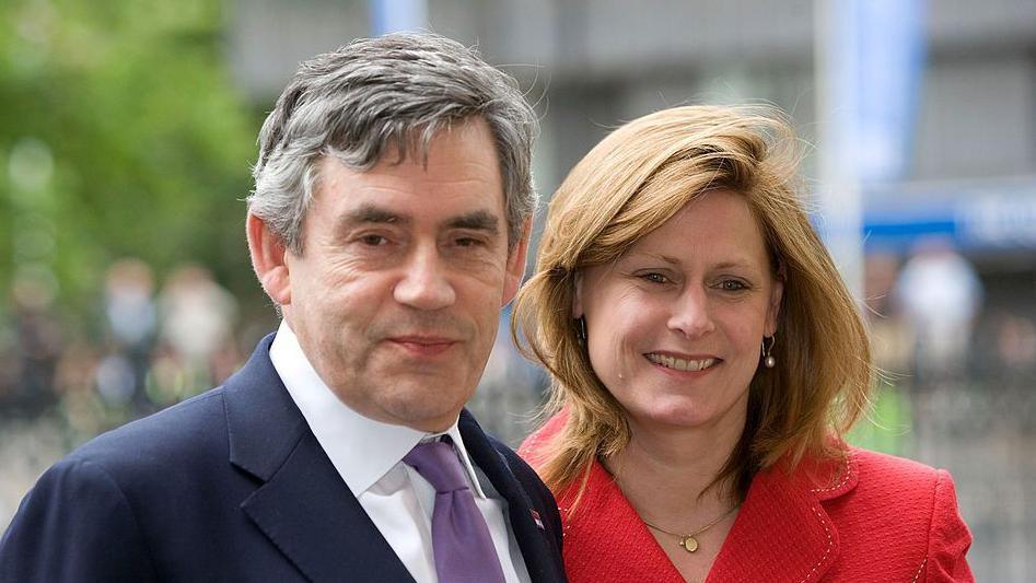 Gordon and Sarah Brown. He has grey hair and is looking at the camera. he is wearing a blue suit with a white shirt and purple tie. Sarah is looking off camera and is wearing a red jacket.