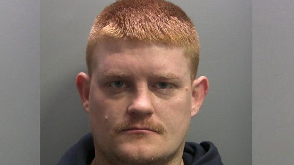 Mugshot of Brian Light. He has short bright ginger hair and has a small ginger moustache. He has blue eyes and is wearing a navy hoodie. 
