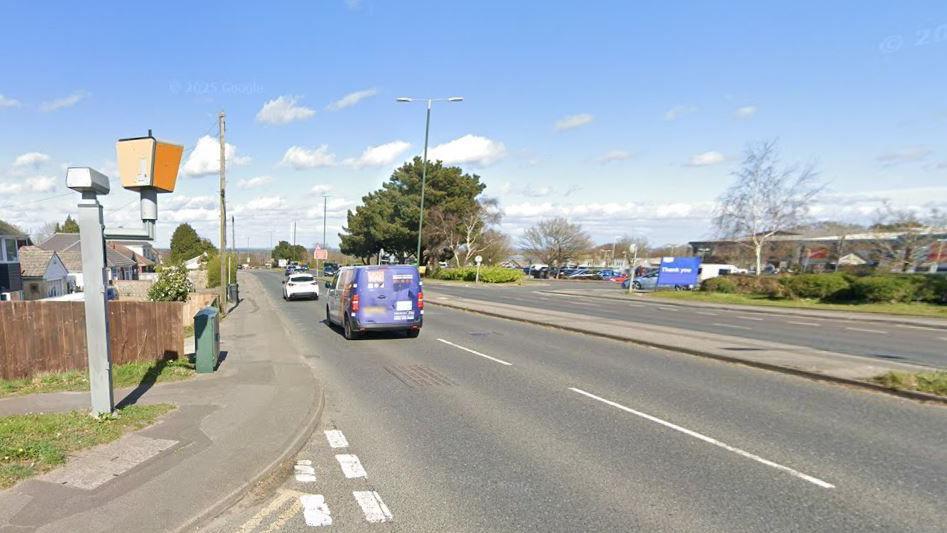 The back of a speed camera can be seen on the left aimed towards a dual carriageway that has a van and car driving on it. A retail park can be seen in the background.