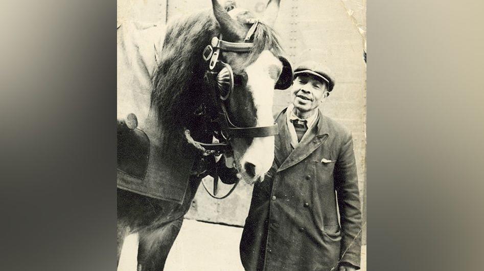 Black and white still of carter Dick Benson dressed in a coat, scarf and cap smiling as he stands next to a horse holding the reins.