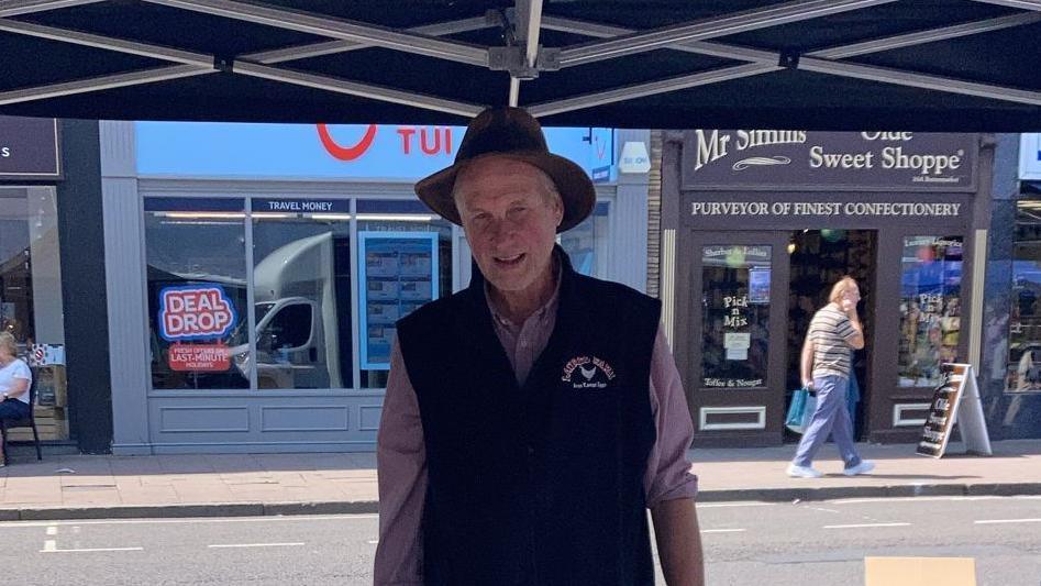 A man standing behind a stall in a town centre. He is wearing a hat and a gilet over a shirt. He is looking into the camera.