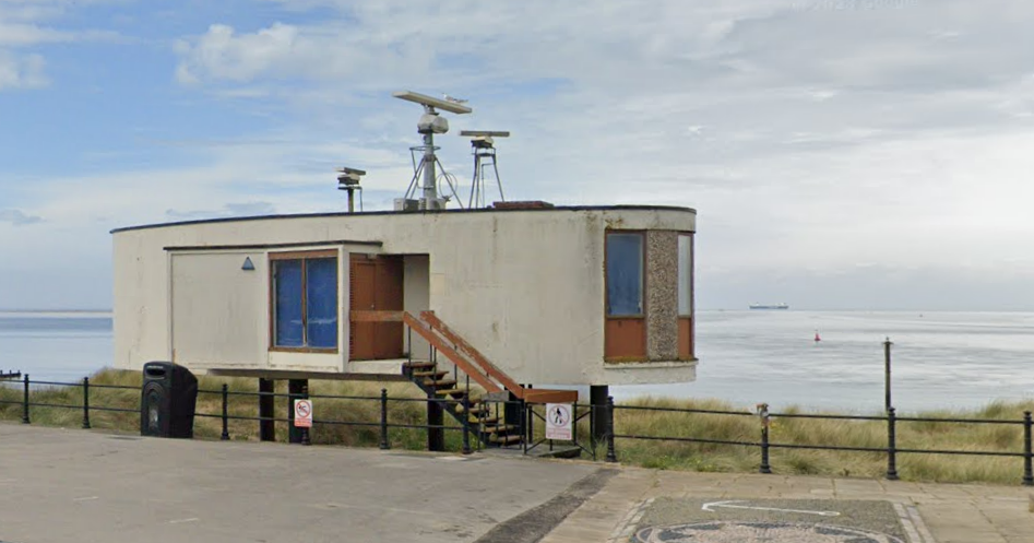 A photo shows the current, "dilapidated" condition of the radar station. While there is no evidence of any structural problems, the building is weathered and the finish is peeling from the short staircase that leads up to the door of the elliptical, single-storey building that stands on stilts on the beach, just behind the promenade railings.