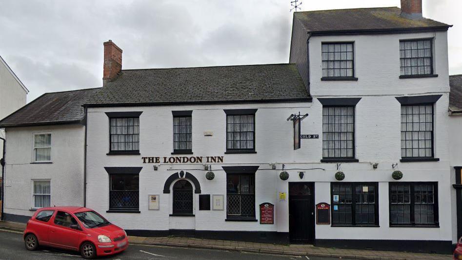 Google Street View image of the London Inn in Ottery St Mary. It is a white brick building with a red car outside on the road. It has several sash windows with black frames. It has a black/grey tiled roof.
