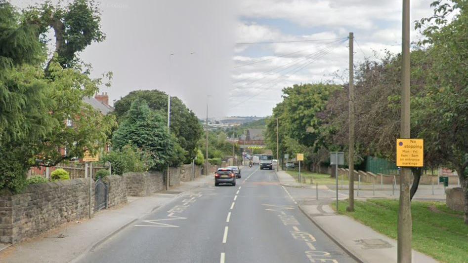 A road with walls and houses on the left hand side. On the right, trees and lampposts. A bridge in the distance.