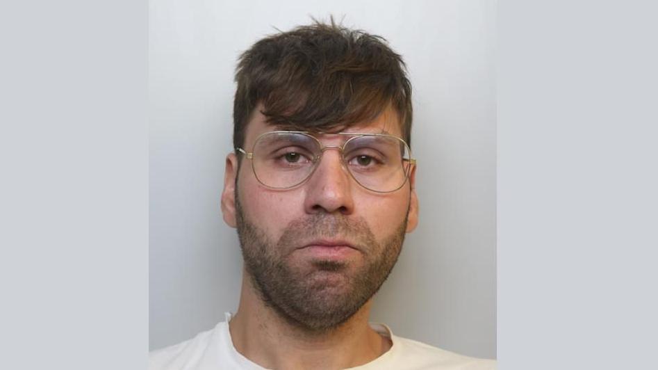 A grumpy-looking wearing glasses man stares at the camera in a police mugshot. He has thick stubble and is wearing a white T-shirt