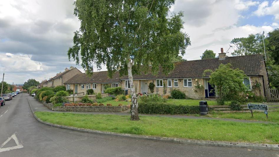 A residential street, with Bennett Place street sign on a grass verge and a row of terraced bungalows with small front gardens behind. Two-storey houses can be seen on the right-hand-side of the street further down, with cars parked on the left.