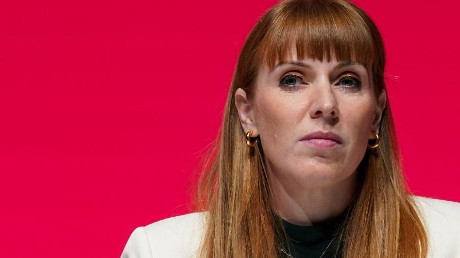 Angela Rayner who has long, ginger hair and is wearing a white suit and gold hooped earrings, looks to her right in front of a red background
