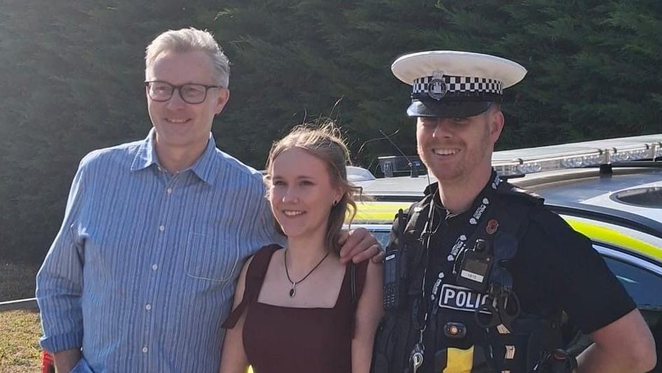Evie stands in between her father and PC Burke in front of a police car. PC Burke wears his police uniform, Evie wears a burgundy prom dress and her father wears a blue shirt and has short grey hair. 