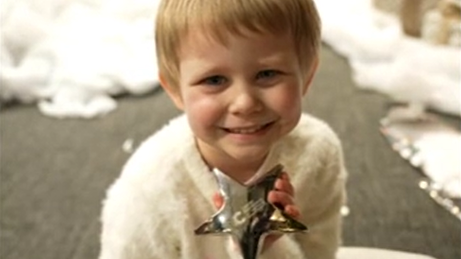 A little girl with short dark blonde hair, wearing a white top, holds a silver star while looking up at the camera and smiling.