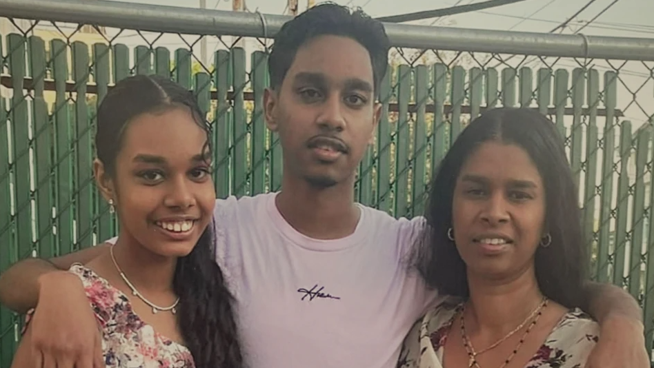 Shawn has his arms around his mother and sister. He is wearing a pink T-shirt. His sister and mother have floral tops on. They are stood in front of a green fence.