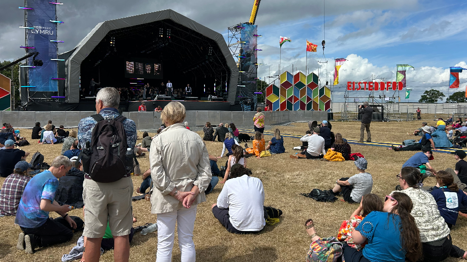 Lots of people sat down on the grass watching a performance on the main stage. On the right, next to the stage, there is a large display of colourful diamonds with flags coming out on the top of it. The red Eisteddfod sign can be seen in the right of the image.