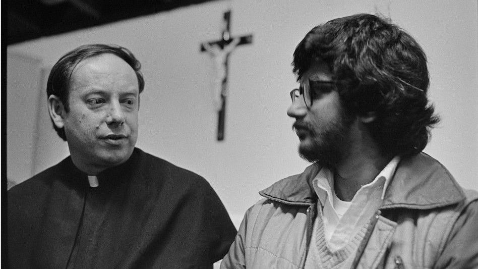 Father John Methuen is sitting in the church next to Viraj. Behind them on the wall is a cross with Jesus Christ. The men are both looking at each other. 