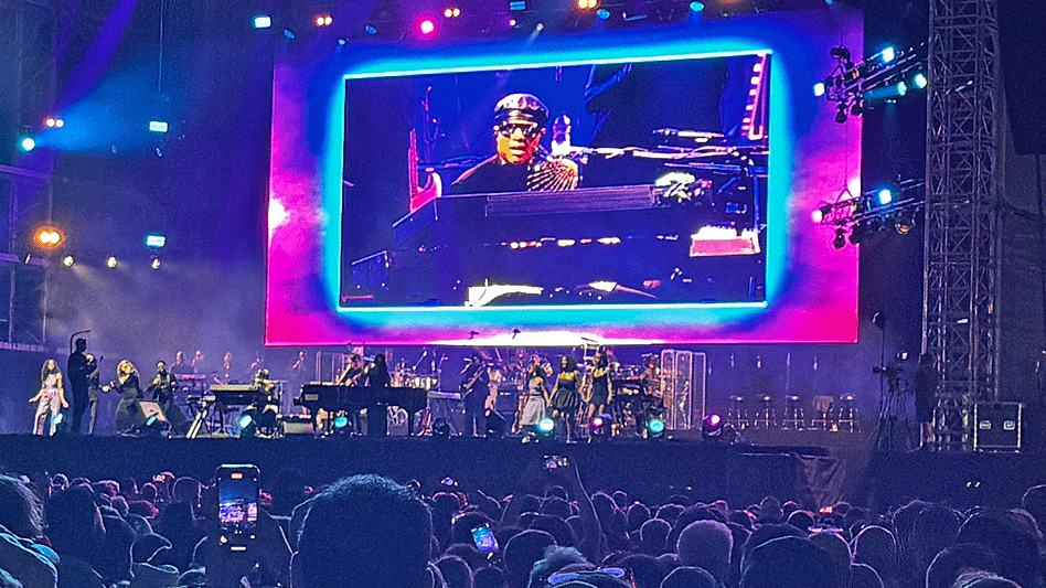Giant live screen showing Stevie Wonder playing a piano and performing on stage in Cardiff, wearing his trademark glasses and a cap