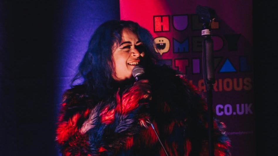 A young woman with dyed-blue hair wearing a red and black furry jacket talking into a microphone on stage.