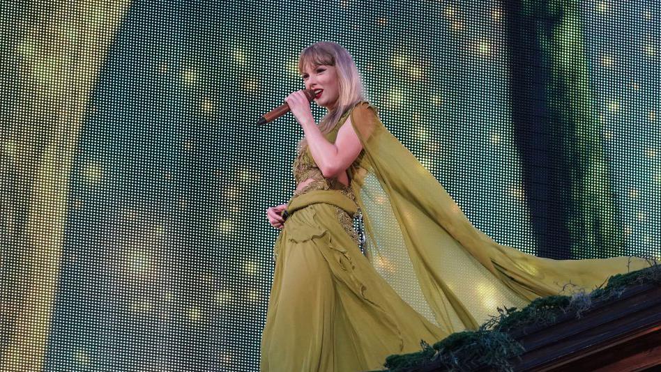 Taylor Swift performs onstage in a green dress performing at one of her recent Era's Tour shows 