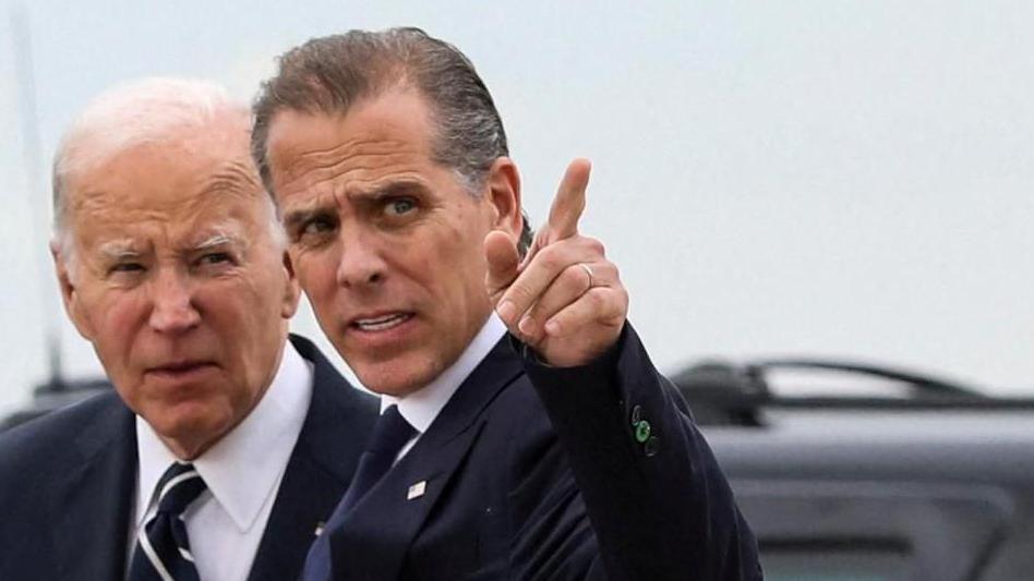 US President Joe Biden stands with his son Hunter Biden, who is pointing a finger at something off frame. They are both wearing navy suits