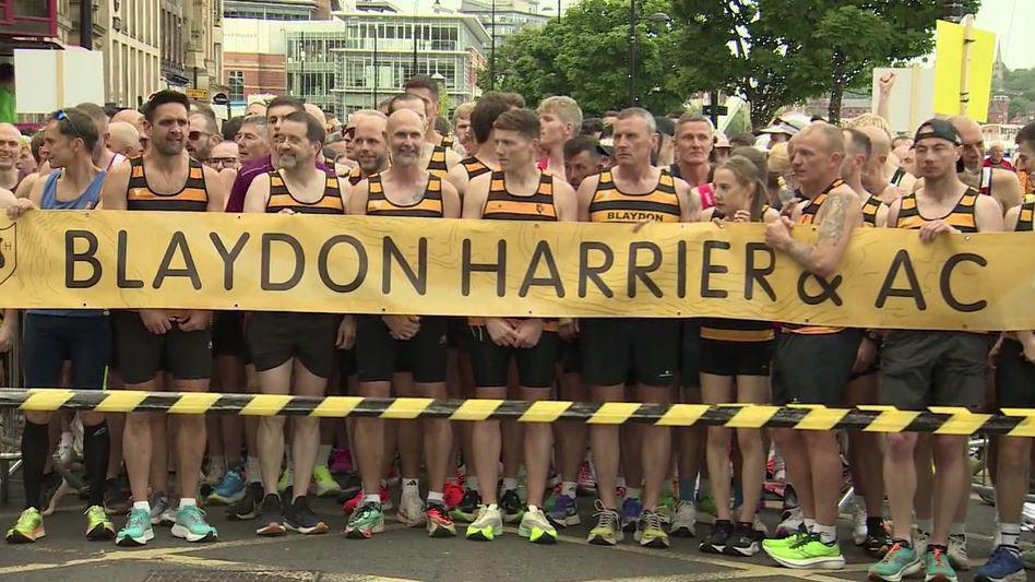 The start line of the Blaydon Race. Thousands of runners line up on Newcastle's Quayside. Dozens of people at the front are wearing yellow and black running tops - and standing behind a paper barrier which reads: "Blaydon Harrier & AC"