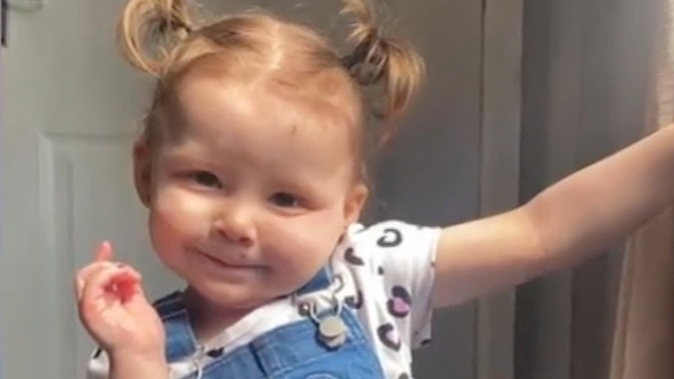 A two year old girl wearing a white t-shirt and blue dungarees stands with one arm raised. She is smiling. Her hair is in high bunches.