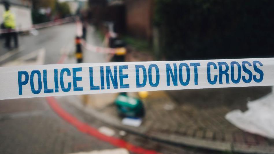 A police tape covers a crime scene with a blurred background of emergency services seen