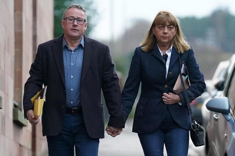 Stuart and Linda Allan walking down a road hand in hand towards the camera. He is wearing blue jeans, a blue shirt and a dark jacket and she she is wearing blue jeans and a blue jacket over a white shirt. She has a handbag over her shoulder and they are both carrying folders. 