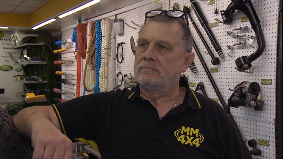 A man with short greying hair and a beard with glasses and a black polo top in a workshop with tools on the wall, resting his hand on a large tyre