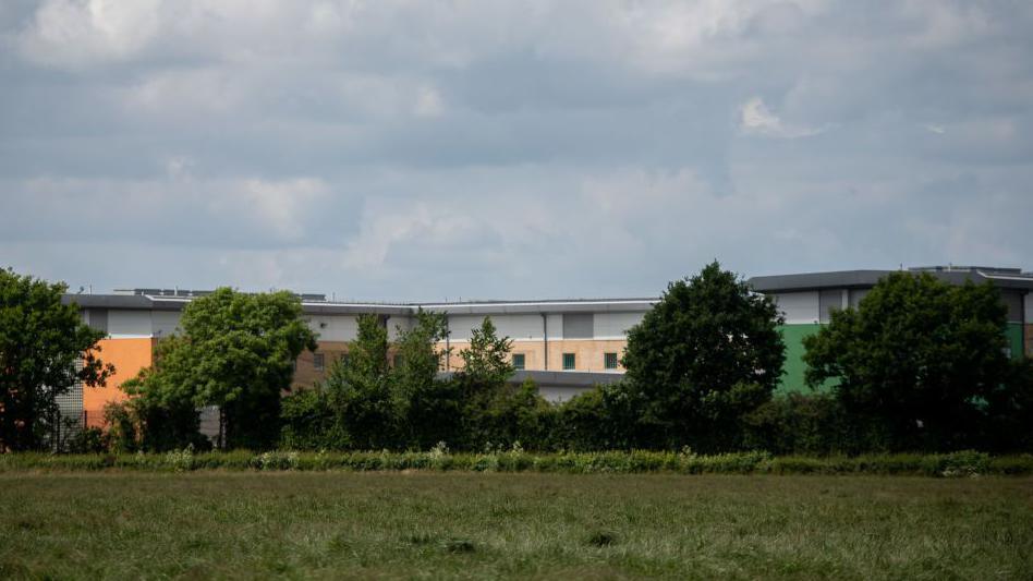 Brook House is seen from over a field with trees partially blocking the view. It is a low modern building with brightly coloured cladding on the lower parts