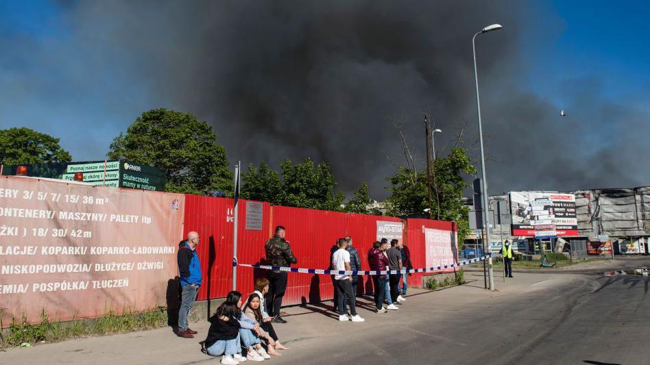 People waiting outside the Marywilska shopping centre as a massive cloud of black smoke comes out of it.
