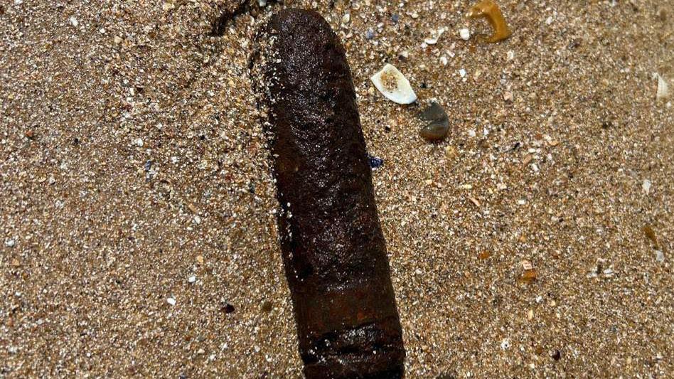 The shell lying flat on the sand. It is unclear how large it is, but it is brown and very rusted.