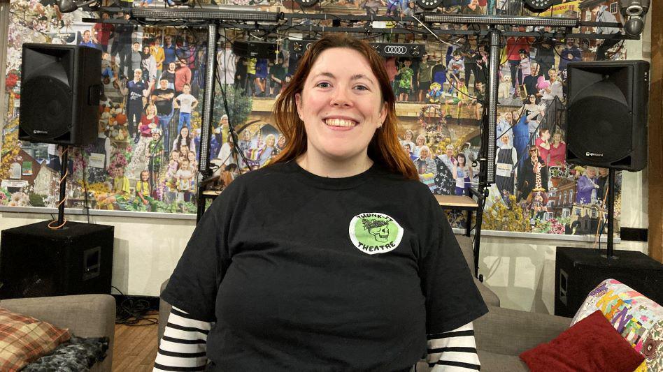 A smiling Becky Lennon wears a black short-sleeved t-shirt with a Thunk-It Theatre logo over a black and white striped long sleeve top. She is standing in front of some theatre equipment and a wall covered in lots of images of people.