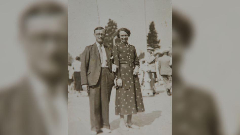A sepia-tone image of a man and a woman. He wears dark trousers and blazer, she wears a high necked dark dress with a light pattern.