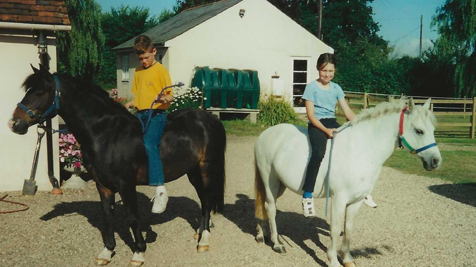 A boy aged about 11 in a yellow T-shirt and blue jeans looks down while sitting on a brown horse, while beside him a girl aged about nine in a pale blue T-shirt and black trousers smiles at the camera while sitting on a white horse.