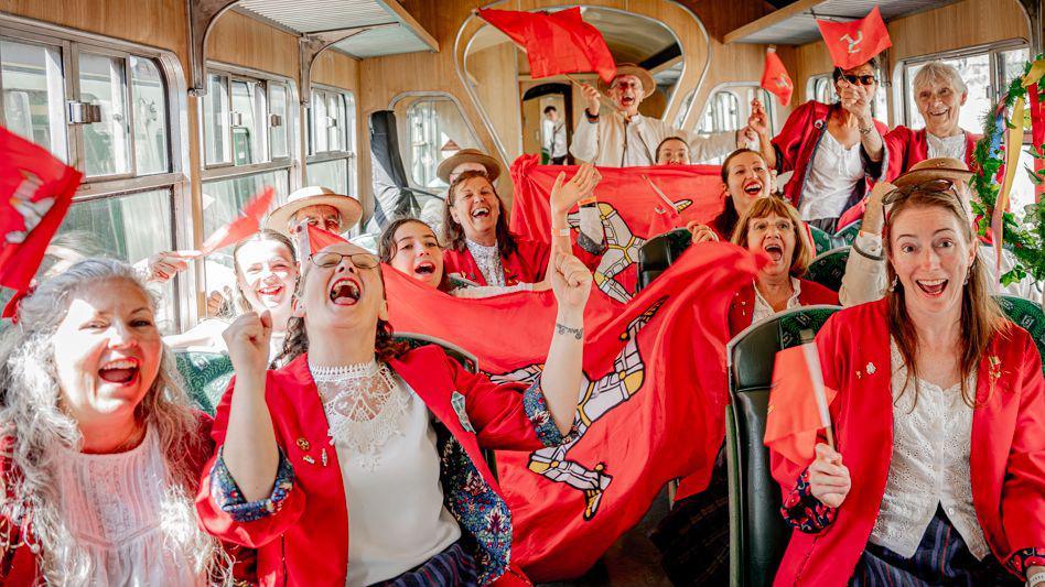 The group sit on a train in their costumes waving red Manx flags and cheer.