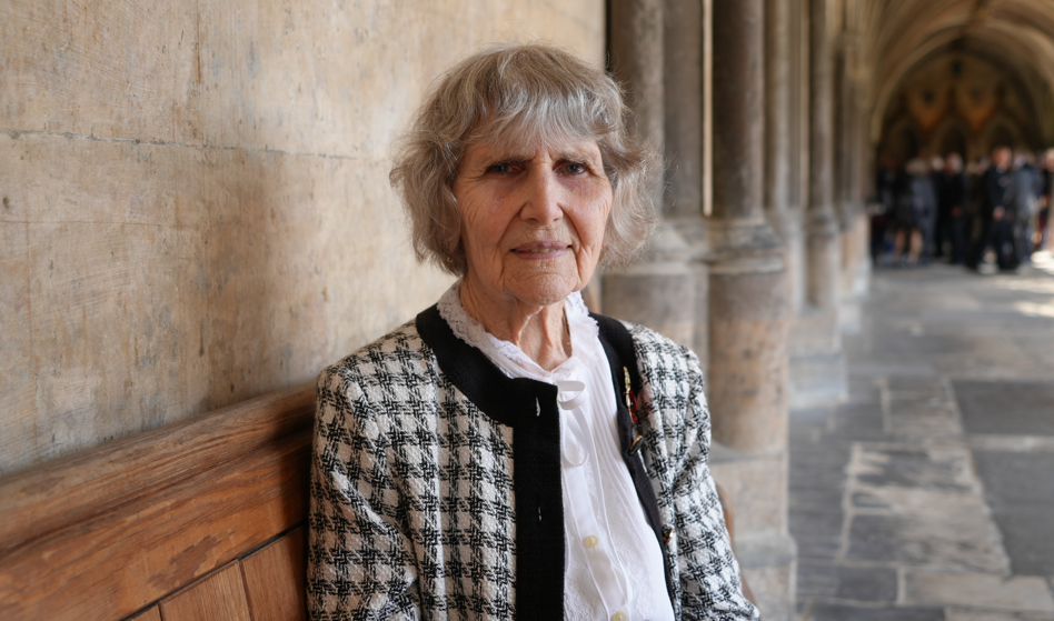 Olga Henderson, with grey hair in a short bob style, sits on a wooden bench in a cathedral. She is wearing a black and white chequered jacket and white blouse  
