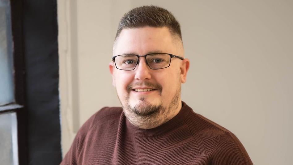 Steve Pressley with short dark brown hair and slight beard, with dark-framed glasses, smiling at the camera and wearing a brown sweat shirt. There is a white wall behind him and window with brown curtain to his right.
