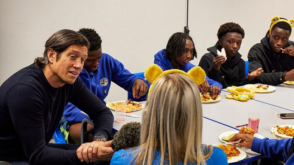 BBC broadcaster Vernon Kay interviewing a youth leader at the youth centre with long blond hair and wearing Children in Need headband while other young people watch on as they eat food  