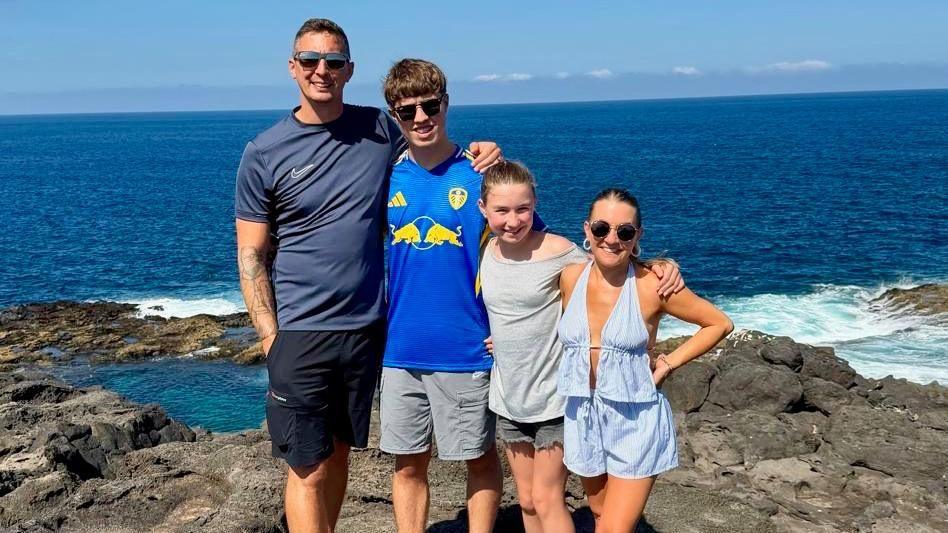 Four people are standing together on a rocky coastline with the ocean in the background. The group is dressed in casual summer clothing, including shorts and light tops. Waves are visible crashing against the rocks behind them, and the horizon shows a clear blue sky with scattered clouds. The setting suggests a scenic seaside location.