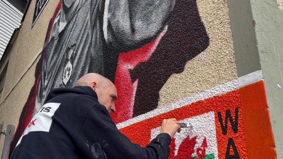 Paul Curtis painting the mural in Wrexham: he is using a brush to apply white paint to the Wales flag, which sits on an orange banner below the image of Joey Jones. The artist wears a dark-coloured hoodie.