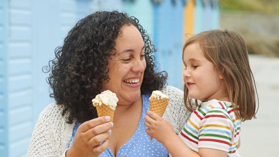 A woman is holding a female child as they eat ice cream. The woman has black curly hair and the child has brown long hair. They are smiling in the picture. the woman is wearing a blue top with white polka dots, and a white cardigan, and the child has a white top with coloured stripes on it.
