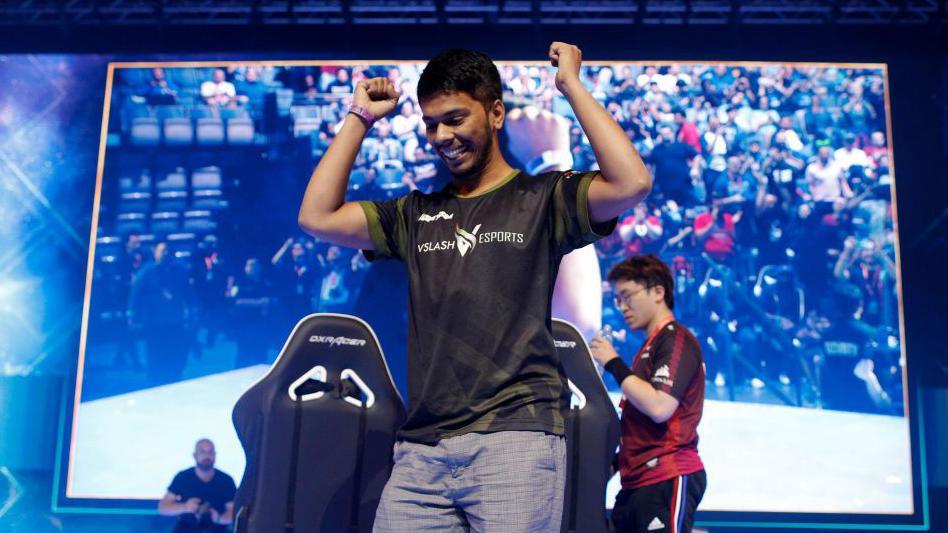 A young man stands in front of a giant screen, smiling with his arms held up in a victory pose. Two gaming chairs, facing the screen, are just behind him. 