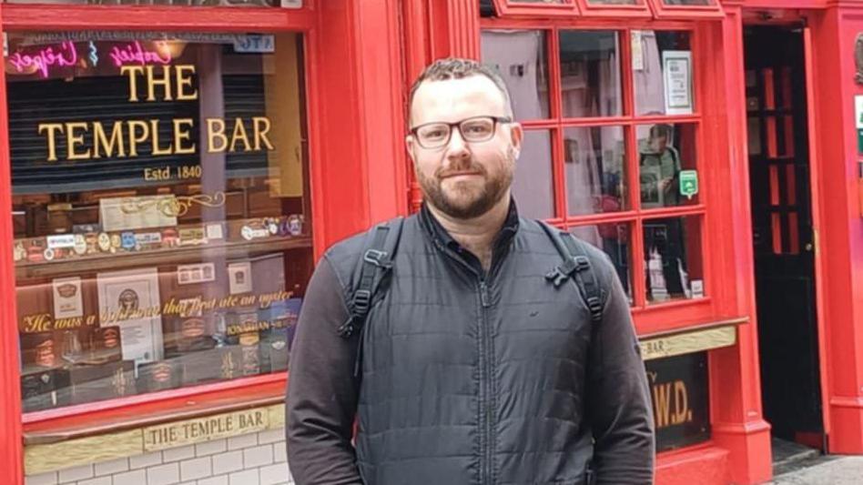 Chris Williams pictured outside The Temple Bar in Dublin, Ireland. He has short dark hair, facial hair and glasses. He is wearing a black coat. He is looking directly at the camera and smiling.