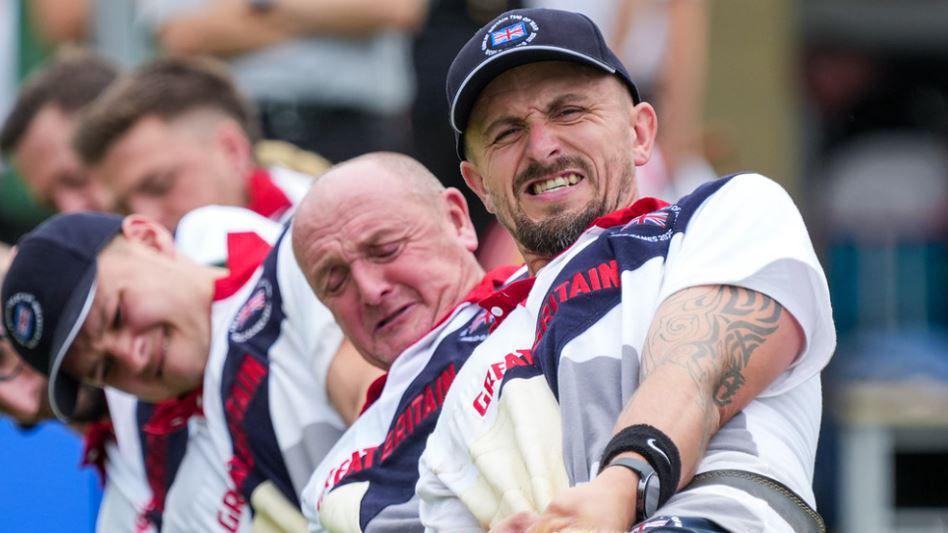 Joe Birch and four of his team members behind him pulling on a rope that is not in the shot. Joe has tattoos and is wearing a GB team cap with union jack emblem. There is strain showing in their faces, some with their eyes screwed up and their faces going red with the effort. 