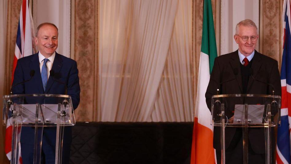 Micheál Martin and Hilary Benn smile off camera as they stand behind separate podiums. Mr Martin is wearing a dark suit, white shirt and blue tie and Mr Benn is wearing a dark suit, dark jumper, blue shirt and red tie. The Union Jack and Irish flags are hanging behind them.