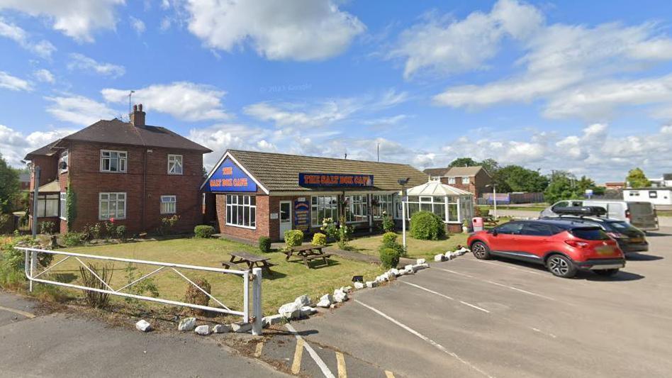 General view of the Salt Box cafe. It has some landscaping at the front along with some parked cares. 