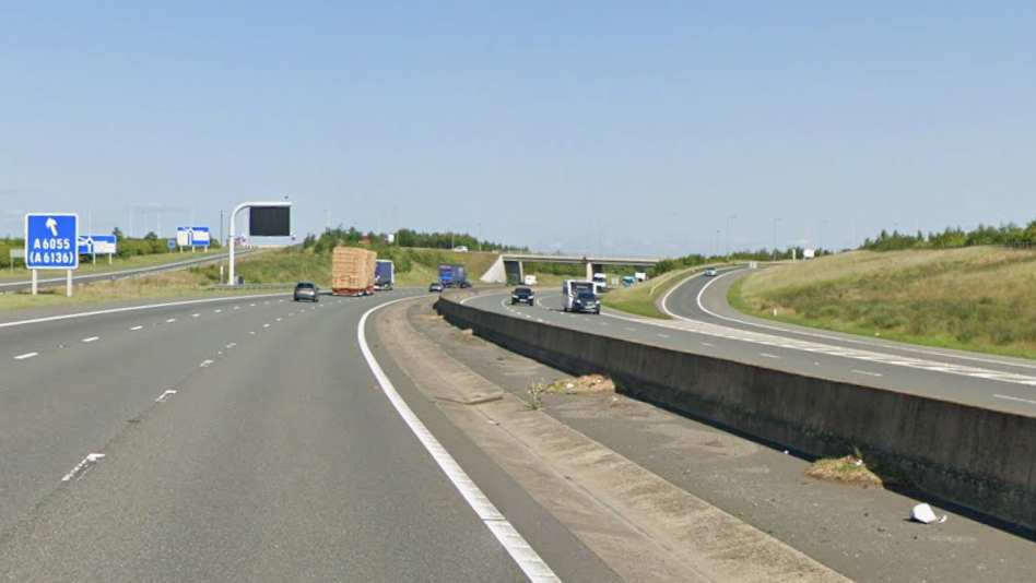 A multi-lane motorway with light traffic under a clear blue sky. Vehicles, including cars and a trailer, are traveling on the left side of the road. A large blue road sign on the left indicates the A6055 (A6136) exit.