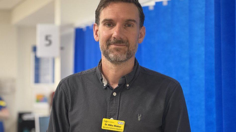 A doctor with dark hair and a beard is wearing a yellow name badge. Blue curtains are behind him.
